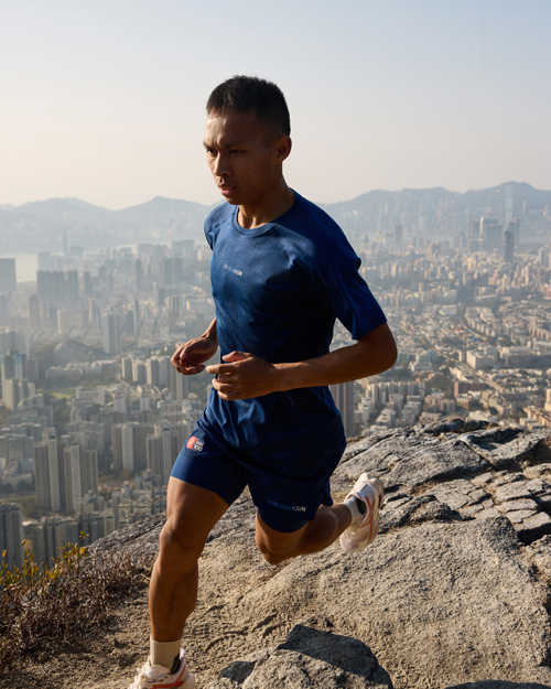 Elite trail runner wearing ANTA GUANJUN 100% Merino wool T-shirt and shorts, jumping across rocky terrain above Hong Kong at sunrise